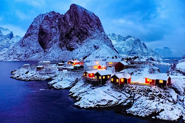 Sneeuwbedekte houten huizen met verlichte ramen aan een rotsachtige kustlijn, tegen een achtergrond van torenhoge, ruige bergen onder een sombere, blauwe lucht.