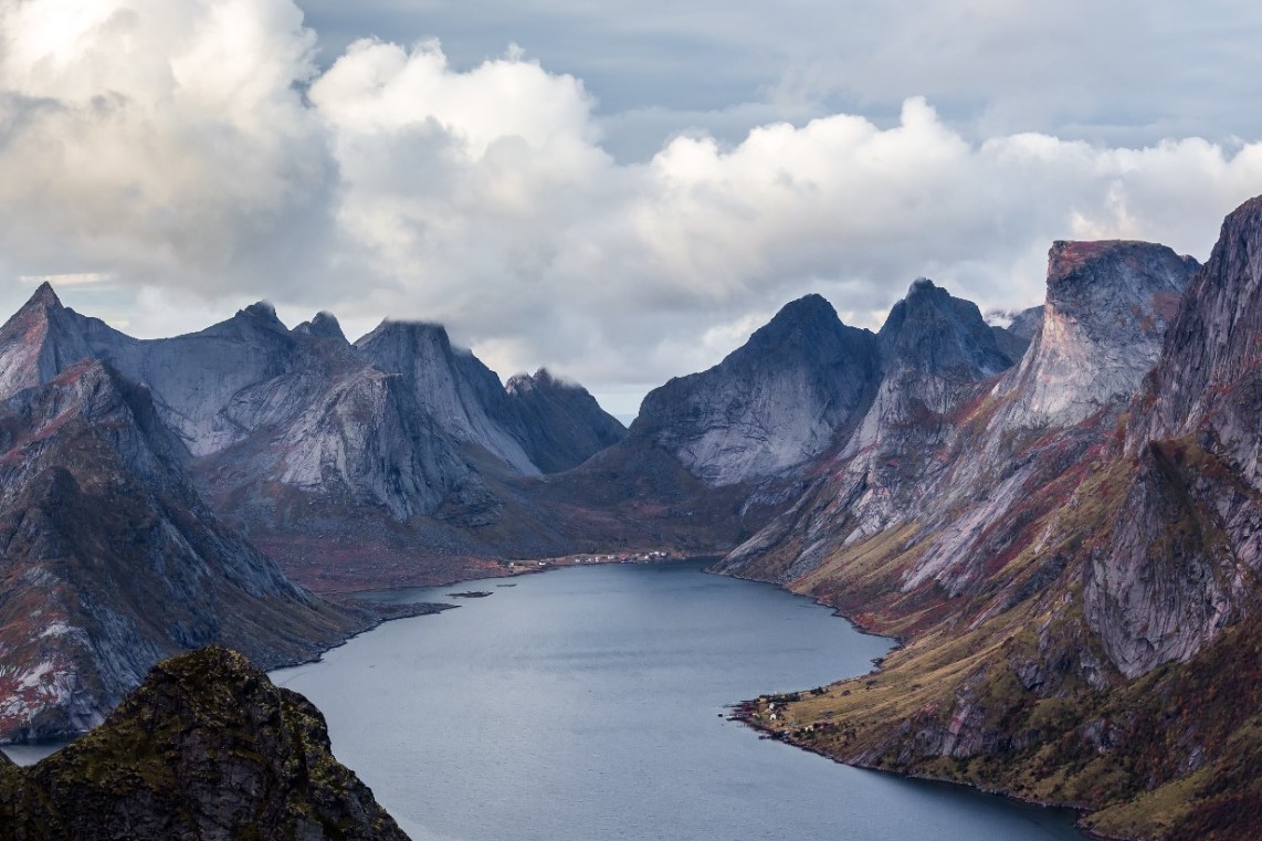 Bergachtig landschap met grijze wolken, een kalm waterlichaam dat door rotsachtige pieken snijdt en een fjordachtige omgeving creëert. De bergen zijn steil, met groene en roodachtige tinten langs de hellingen.
