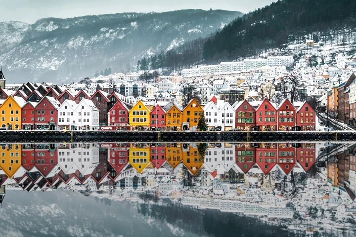 Kleurrijke huizen weerspiegeld in kalm water in een besneeuwd landschap, met rode, gele en oranje gebouwen langs de waterkant en sneeuwbedekte heuvels en bomen op de achtergrond.