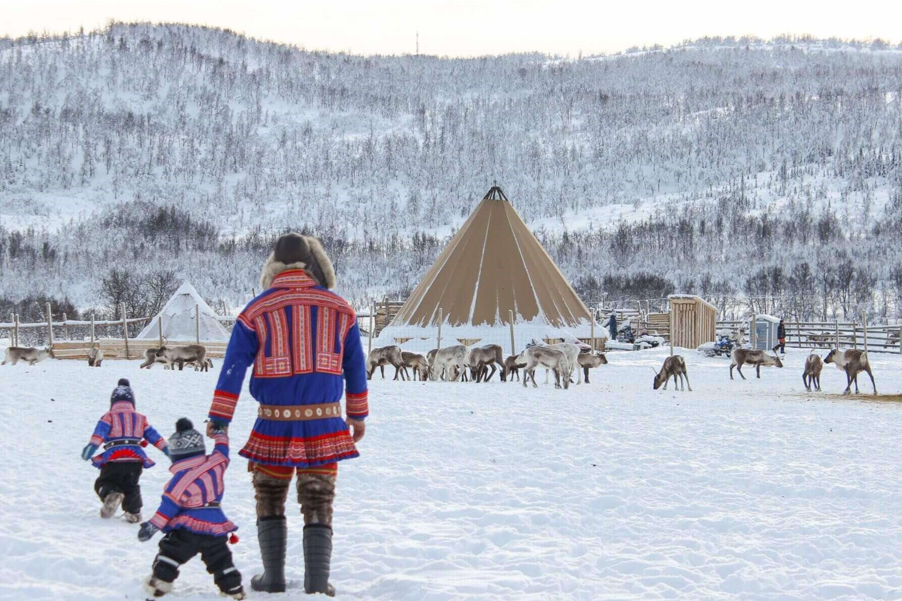 Drie mensen in traditionele kleding lopen door de sneeuw naar een kegelvormige tent en rendieren, met besneeuwde heuvels op de achtergrond.