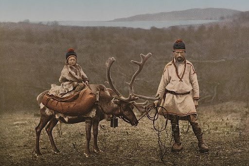 Een persoon in traditionele kledij staat naast een rendier met een kind op zijn rug, beiden dragen wollen mutsen. Op de achtergrond een uitgestrekt landschap met water en heuvels.